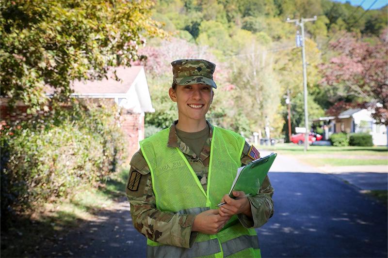 Jessica Bowling in National Guard uniform holding a clipboard and smiling for a portrait while standing on a street in a neighborhood in Waynesville, North Carolina.