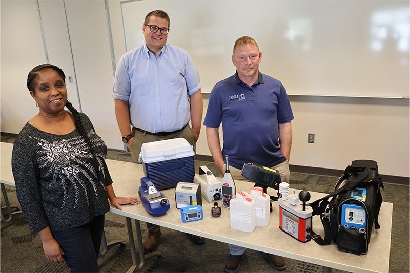 Dave Catalano, Greg Williams and Kim Haley posing for a photo by table full of safety equipment