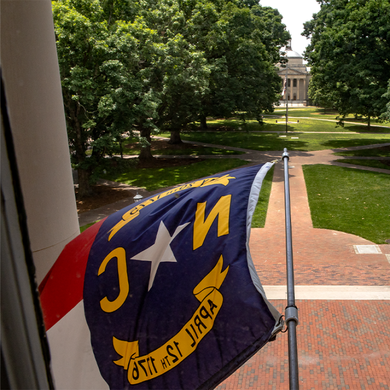 North Carolina state flag flying from the the exterior of South Building on the campus of UNC-Chapel Hill. Polk Place, known colloquially as the Quad, is seen in the background.