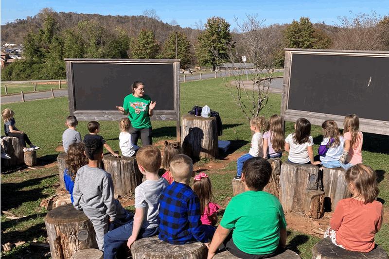 Elementary aged children listening to an outdoor science lesson on a grass field.